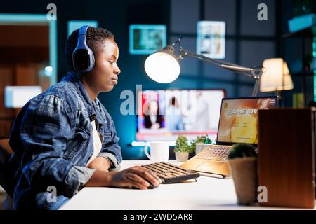 Black woman with headphones sits in front of wireless computer, which is showing game over. Female player is staring at her digital laptop screen while wearing a headset and losing a game. Stock Photo
