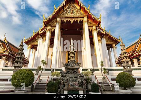 Entrance to Wat Suthat Temple in Bangkok, thailand. Ornamental garden in front. Other buildings on either side; blue sky and clouds above. Stock Photo