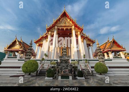 Entrance to Wat Suthat Temple in Bangkok, thailand. Ornamental garden in front. Other buildings on either side; blue sky and clouds above. Stock Photo