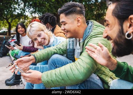 A group of diverse friends laugh and share content on a smartphone while sitting together in an urban outdoor setting, portraying modern connectivity Stock Photo