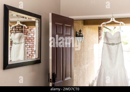 Elegant white wedding gown hanging gracefully on a wall near an open doorway Stock Photo