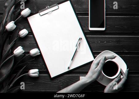 White tulips on a wooden table with an empty paper tablet and smartphone and a Cup of coffee in women's hands. Flat lay, top view composition with cop Stock Photo