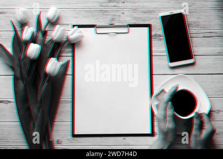 A white bouquet of tulips on a wooden background with a coffee Cup in women's hands, a smartphone and an empty paper tablet. Black and white photo wit Stock Photo