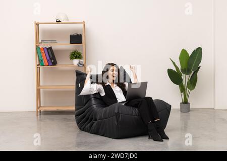 Happy businesswoman showing thumps up sitting on chair in front of gray wall at office Stock Photo