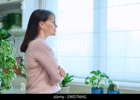 Dreamy, thoughtful middle-aged woman looking out the window at home Stock Photo
