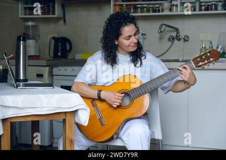 young Venezuelan woman at home sitting happy smiling, with her laptop on table and in online guitar class, rehearsing learning to play new songs. musi Stock Photo