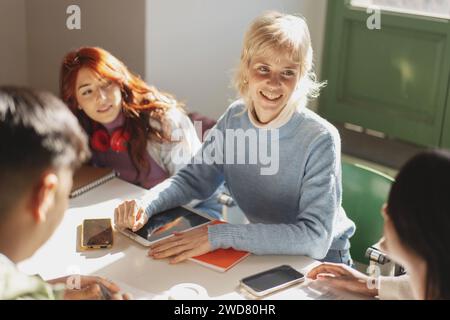 A group of cheerful college students collaboratively using tablets and smartphones for their study session in a bright classroom, showcasing interacti Stock Photo