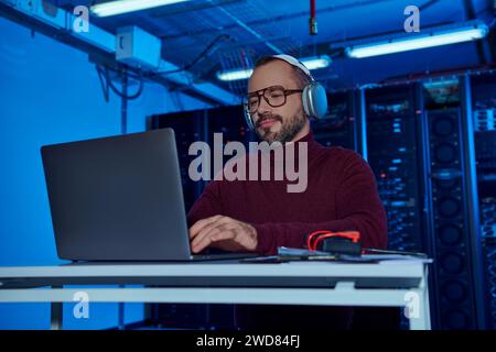 cheerful attractive specialist in turtleneck with headphones working on his laptop, data center Stock Photo