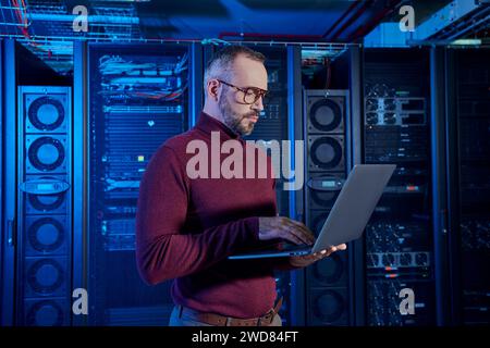attractive bearded specialist in turtleneck working attentively at his laptop, data center Stock Photo