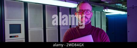 pensive specialist in turtleneck holding laptop and looking at camera, data center, banner Stock Photo