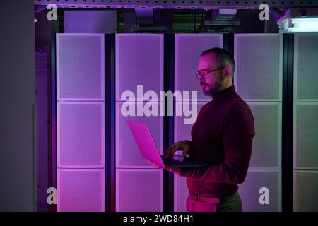 handsome jolly professional in turtleneck with glasses and beard working on laptop, data center Stock Photo