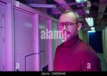 cheerful dedicated specialist in cozy turtleneck with glasses smiling at camera, data center Stock Photo