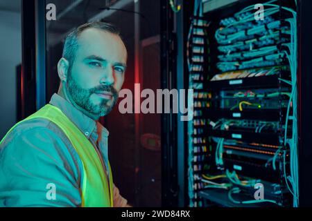 attractive pensive man with beard in safety vest inspecting data center and looking at camera Stock Photo