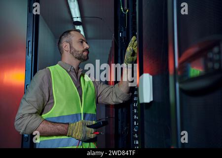 pensive specialist in safety vest and gloves using detector during inspection of data center Stock Photo