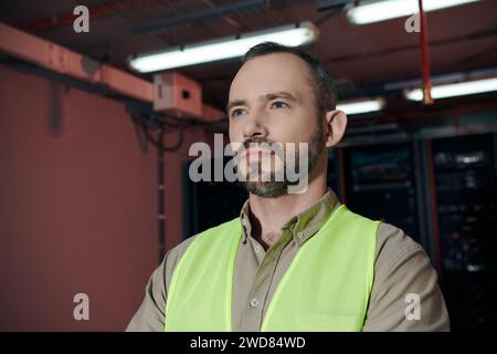attractive pensive specialist in safety vest posing and looking away while working in data center Stock Photo