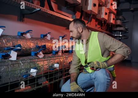 good looking pensive technician in safety gloves and vest squatting while working in data center Stock Photo