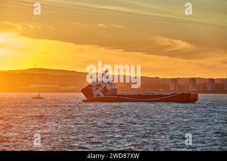 Platform supply vessel sails in port, during sunset sky and city coastline on the horizon. Stock Photo