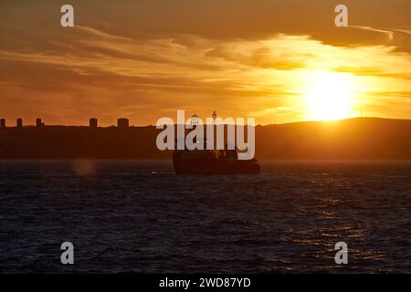 A ship sails through the waters of city coastline, during golden sunset, with sun on the horizon and a ray of sunlight breaks through the cloud. Stock Photo