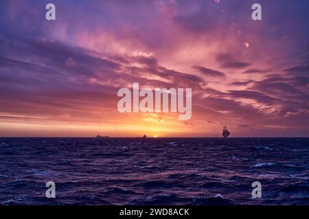 Ocean landscape with colourful pink, orange, blue and red sky, and a silhouette of a jack up drilling rig on the horizon. Stock Photo