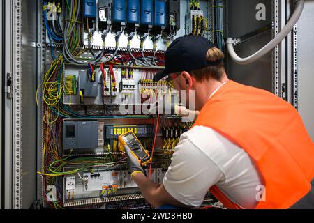 an electrician in a cap, yellow glasses and an orange vest measures electric current with a digital multimeter on a distribution box. Stock Photo