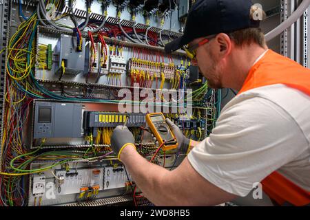 an electrician in a cap, yellow glasses and an orange vest measures electric current with a digital multimeter on a distribution box. Stock Photo