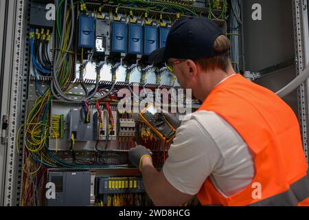 an electrician in a cap, yellow glasses and an orange vest measures electric current with a digital multimeter on a distribution box. Stock Photo