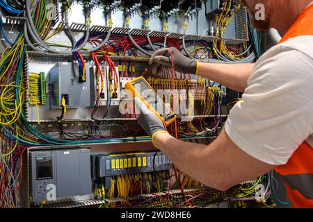 an electrician in a cap, yellow glasses and an orange vest measures electric current with a digital multimeter on a distribution box. Stock Photo
