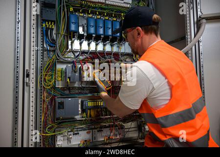 an electrician in a cap, yellow glasses and an orange vest measures electric current with a digital multimeter on a distribution box. Stock Photo
