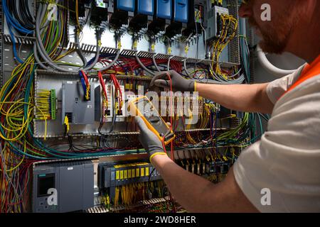 an electrician in a cap, yellow glasses and an orange vest measures electric current with a digital multimeter on a distribution box. Stock Photo