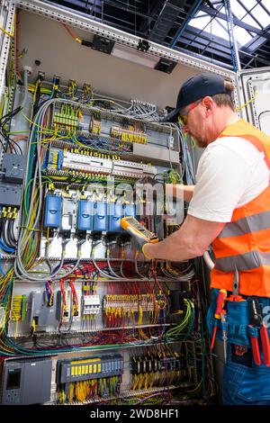 an electrician in a cap, yellow glasses and an orange vest measures electric current with a digital multimeter on a distribution box. Stock Photo