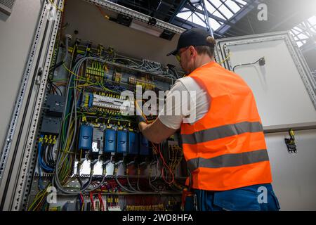 an electrician in a cap, yellow glasses and an orange vest measures electric current with a digital multimeter on a distribution box. Stock Photo