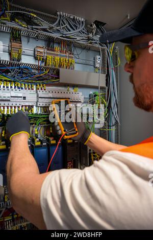 an electrician in a cap, yellow glasses and an orange vest measures electric current with a digital multimeter on a distribution box. Stock Photo