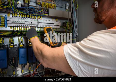 an electrician in a cap, yellow glasses and an orange vest measures electric current with a digital multimeter on a distribution box. Stock Photo