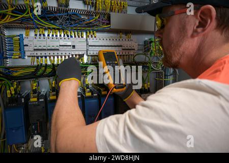 an electrician in a cap, yellow glasses and an orange vest measures electric current with a digital multimeter on a distribution box. Stock Photo