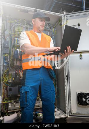an electrician in a cap and orange vest installs a large electrical substation using a computer. Stock Photo