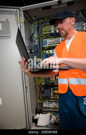 an electrician in a cap and orange vest installs a large electrical substation using a computer. Stock Photo