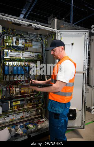 an electrician in a cap and orange vest installs a large electrical substation using a computer. Stock Photo
