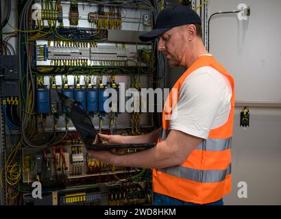 an electrician in a cap and orange vest installs a large electrical substation using a computer. Stock Photo