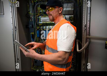 an electrician in a cap and orange vest installs a large electrical substation using a tablet. Stock Photo