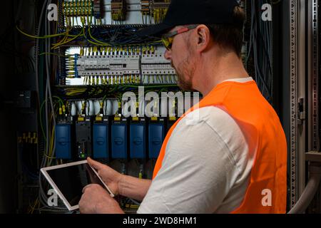an electrician in a cap and orange vest installs a large electrical substation using a tablet. Stock Photo