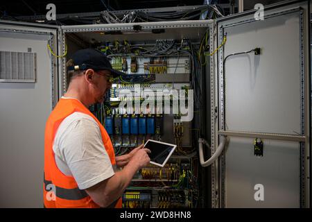 an electrician in a cap and orange vest installs a large electrical substation using a tablet. Stock Photo
