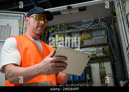an electrician in a cap and orange vest installs a large electrical substation using a tablet. Stock Photo