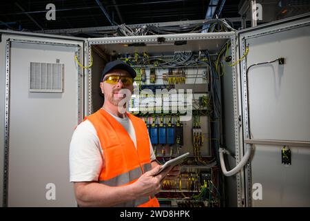 an electrician in a cap and orange vest installs a large electrical substation using a tablet. Stock Photo