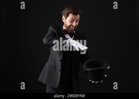 Happy magician showing magic trick with top hat on black background Stock Photo