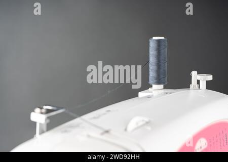 Spool of gray sewing thread stands on top of a white modern sewing machine. Sewing process, bobbin with thread close up Stock Photo