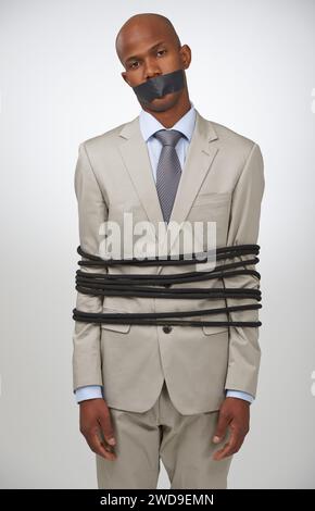 Portrait, business and black man tied with rope, mouth covered with tape and compliance issue on white studio background. African person, employee or Stock Photo