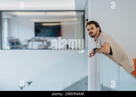 A thoughtful man with a beard leans on a glass partition in a modern office space, looking into the distance. Stock Photo