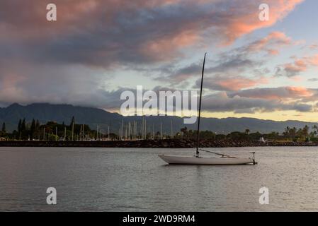 Single boat, in a calm sea, at sunset on the north shore of Oahu, Hawaii. There are dark storm clouds rolling over the hills flanking the beach Stock Photo