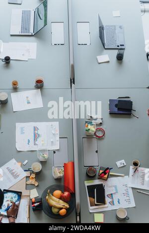 Overhead view of a collaborative workspace featuring two laptops, tablets, office supplies, fruit, and various business charts sprawled on a table. Stock Photo