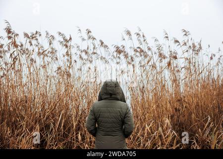 Lonely woman is wearing a hooded coat and standing against reed grass by misty lake. Loneliness concept Stock Photo